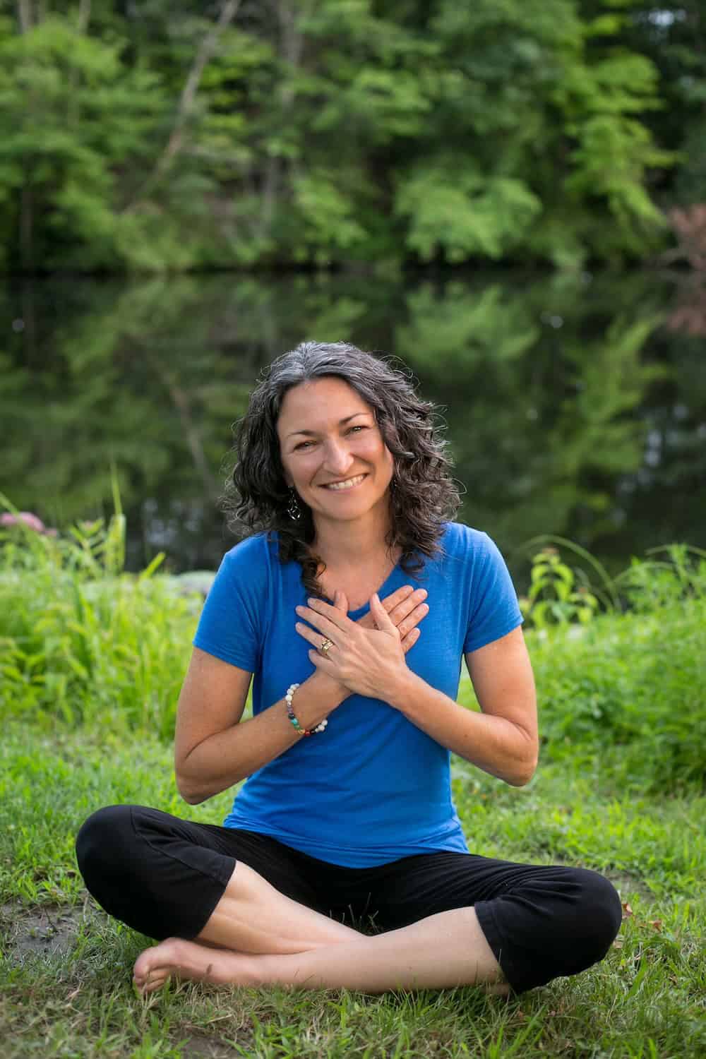 Val Nelson - holistic coach for introverts and sensitive souls - seated in lotus position with hands over her heart and smiling. sitting on the grass by a pond.