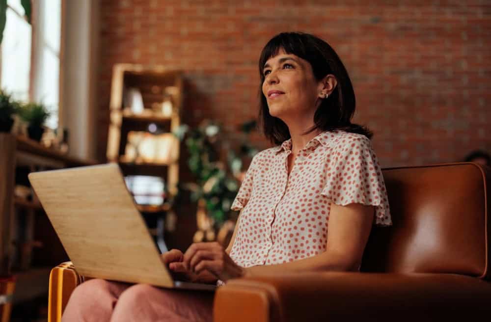 midlife woman writer sitting with laptop in a living room, pondering her writing.